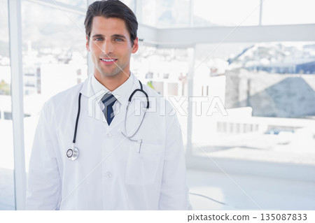 Male doctor standing in clinic by windows holding stethoscope while wearing white lab coat 135087833