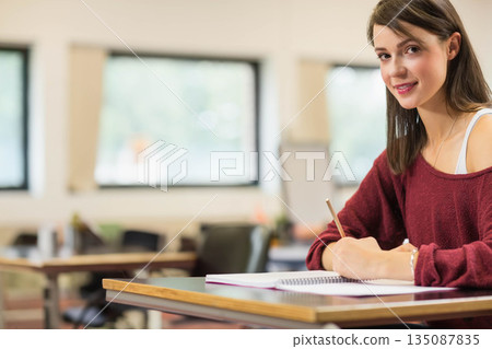 Female student writing in notebook with pencil at desk near windows and flip chart, copy space Female student writing in notebook with pencil at desk near windows and flip chart, copy space 135087835