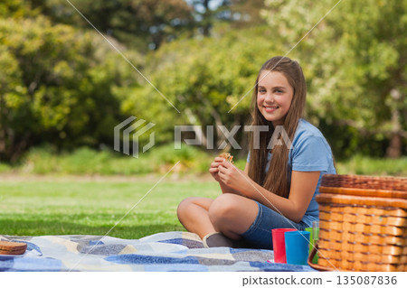 Child female sitting on blanket in park holding sandwich near picnic basket and cups, copy space Child female sitting on blanket in park holding sandwich near picnic basket and cups, copy space 135087836