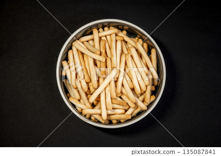 Crispy small breadsticks served on a white glass plate against a black background. Minimalistic food composition highlighting texture, contrast, and simple snack presentation. 135087841