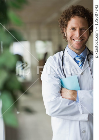 Male doctor standing in hospital hallway holding blue folder and wearing stethoscope, copy space Male doctor standing in hospital hallway holding blue folder and wearing stethoscope, copy space 135087842