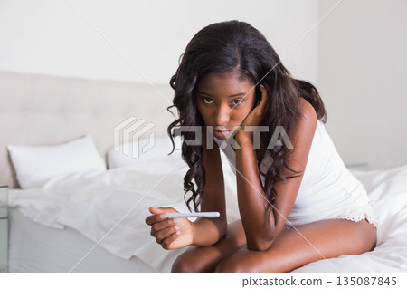 African American woman in sleepwear sitting on bed holding pregnancy test stick looking concerned African American woman in sleepwear sitting on bed holding pregnancy test stick looking concerned 135087845