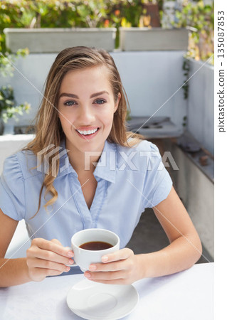 Woman sitting on café patio holding white coffee cup and smiling toward camera on sunny terrace Woman sitting on café patio holding white coffee cup and smiling toward camera on sunny terrace 135087853