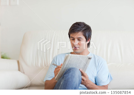 Male sitting on padded sofa in living room, solving puzzle in folded newspaper with ballpoint pen Male sitting on padded sofa in living room, solving puzzle in folded newspaper with ballpoint pen 135087854