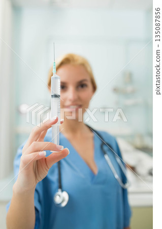 Female nurse wearing blue scrubs with stethoscope holding clear-fluid syringe in exam room Female nurse wearing blue scrubs with stethoscope holding clear-fluid syringe in exam room 135087856