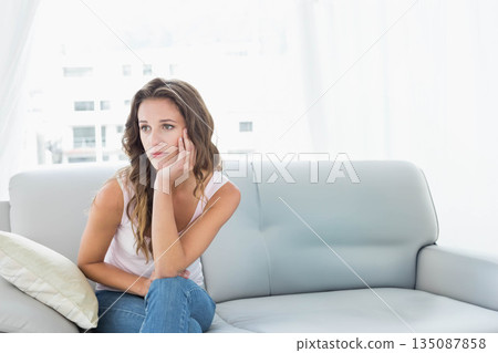 Woman sitting at home on gray leather sofa leaning forward with hand supporting head, copy space 135087858