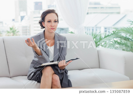 Female professional sitting on white leather sofa in office lounge holding black folder gesturing 135087859