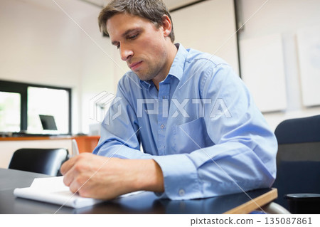 Male student writing notes in notebook at desk in classroom with laptop and projection screen Male student writing notes in notebook at desk in classroom with laptop and projection screen 135087861