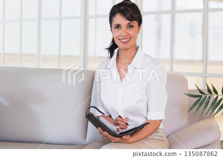 Asian woman writing with silver pen in black planner on sofa in office lounge, copy space 135087862