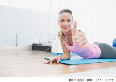 Female holding pose on blue mat in gym sportswear giving thumbs-up with stability ball, copy space 135087863