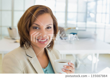 Woman wearing blazer holding white coffee cup smiling at office desk with laptop, copy space 135087864