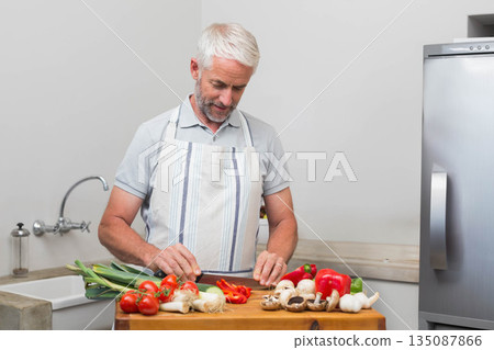 Senior man wearing striped apron chopping red bell peppers on board with chef's knife in kitchen Senior man wearing striped apron chopping red bell peppers on board with chef's knife in kitchen 135087866