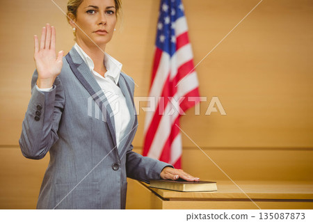 Woman standing in witness stand raising right hand on Bible beside US flag in grey suit Woman standing in witness stand raising right hand on Bible beside US flag in grey suit 135087873