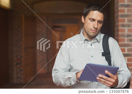 Man standing in brick hallway using tablet and wearing backpack with button-down shirt, copy space 135087876