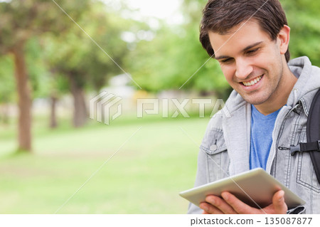 Man standing in grassy park holding tablet with both hands and carrying black backpack, copy space Man standing in grassy park holding tablet with both hands and carrying black backpack, copy space 135087877