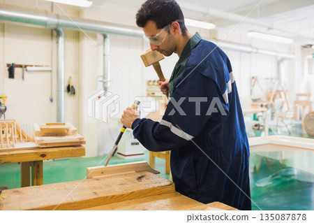 Male woodworker wearing goggles and coverall carving wood block at workbench with mallet and chisel Male woodworker wearing goggles and coverall carving wood block at workbench with mallet and chisel 135087878