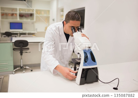 Male researcher leaning over microscope adjusting focus knobs on lab bench with computer 135087882