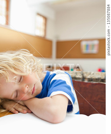 School-age boy resting head on folded arms on wooden desk in classroom with blank notebook paper School-age boy resting head on folded arms on wooden desk in classroom with blank notebook paper 135087884