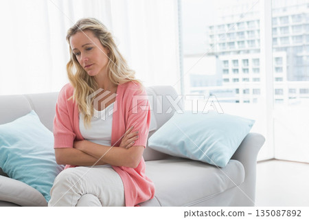 Woman sitting on sofa in living room wearing cardigan, crossing arms, gazing at windows, copy space Woman sitting on sofa in living room wearing cardigan, crossing arms, gazing at windows, copy space 135087892