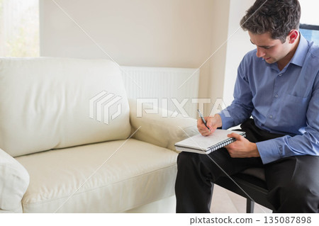 Man in late twenties writing with pen in spiral-bound notebook in home office, copy space 135087898