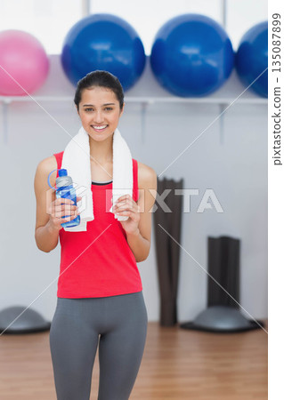 Asian female holding blue water bottle near stability balls and wearing red top in fitness studio 135087899