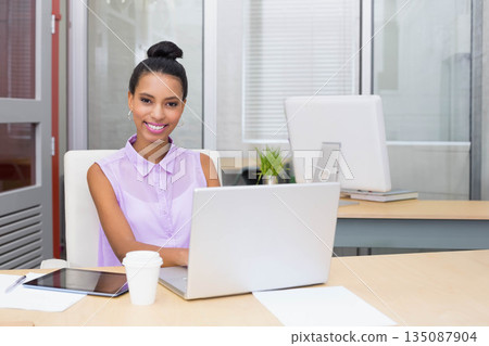 African American woman smiling while using silver laptop at office desk with tablet and coffee cup 135087904