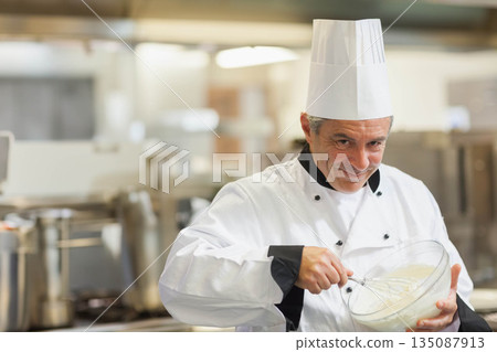 Male chef wearing toque whisking mixture with metal whisk in bowl in restaurant kitchen, copy space 135087913