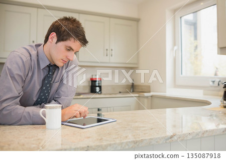 Man wearing business attire leaning on island using tablet beside coffee mug in kitchen, copy space 135087918