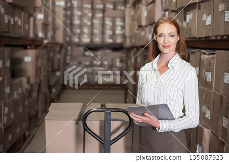 Mid-adult female warehouse supervisor holding clipboard inspecting pallet jack in aisle, copy space 135087923
