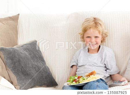 Child boy holding salad plate and TV remote while sitting on sofa at home, copy space 135087925
