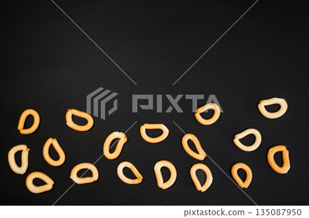 Crispy small dry bread bagels scattered on a black background. Minimalistic snack food composition highlighting simple texture, contrast, and bakery concept. Crispy small dry bread bagels scattered on a black background. Minimalistic snack food composition highlighting simple texture, contrast, and bakery concept. 135087950