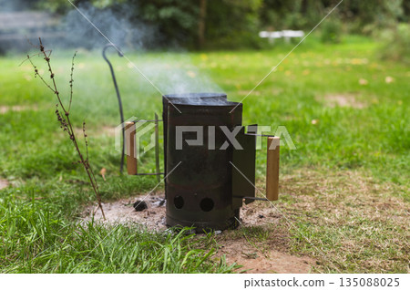 Smoke coming out of a smokestack of a small black smoker grill or barbecue on green background. High quality photo 135088025