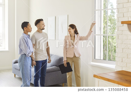 Real estate agent showing new house and window view to a young smiling couple. Real estate agent showing new house and window view to a young smiling couple. 135088458