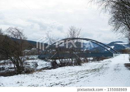 Modern arched bridge over river in winter landscape. Grey metal bridge in snowy countryside with mountains Modern arched bridge over river in winter landscape. Grey metal bridge in snowy countryside with mountains 135088989