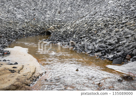 Water moves slowly through a channel filled with rocks and gravel. A drainage pipe is visible in the background. The scene shows a construction site 135089000