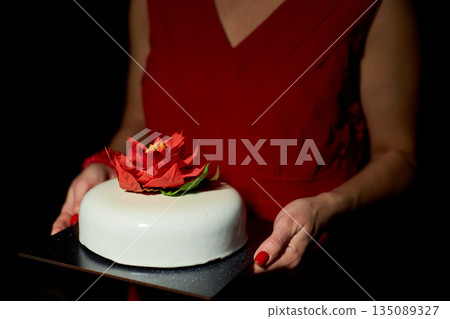 A Person Holding a Beautifully Decorated Cake Adorned With a Vibrant Flower in a Dimly Lit Setting During a Special Celebration 135089327