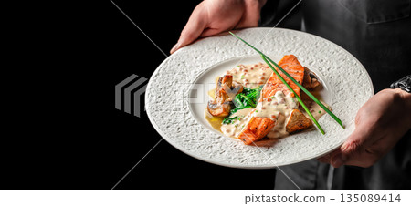 Chef holding a plate of grilled salmon with creamy sauce, spinach, and mushrooms on a white plate. copy space, top view 135089414