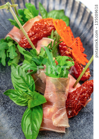 Prosciutto rolls with arugula and sun-dried tomatoes on a dark plate. top view 135089618