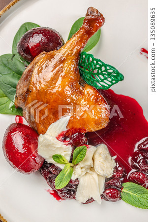 Glazed duck leg served with dark berry sauce, fresh cherries, cream cheese, and vibrant herbs on a white plate 135089635