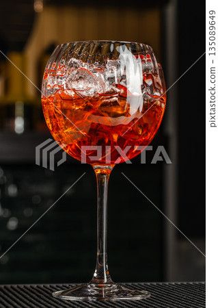 A vibrant cocktail served in a large stemmed glass with clear ice cubes, glowing in warm orange and red tones against a moody bar background A vibrant cocktail served in a large stemmed glass with clear ice cubes, glowing in warm orange and red tones against a moody bar background 135089649