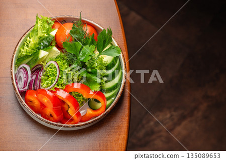 A fresh and colorful vegetable salad featuring sliced cucumbers, red bell peppers, tomatoes, red onions, lettuce, and fragrant herbs on a rustic wooden table A fresh and colorful vegetable salad featuring sliced cucumbers, red bell peppers, tomatoes, red onions, lettuce, and fragrant herbs on a rustic wooden table 135089653