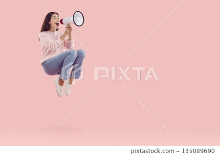 Happy woman yelling through megaphone while jumping on pink copy space background Happy woman yelling through megaphone while jumping on pink copy space background 135089690