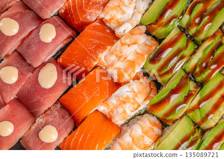 An overhead view of assorted sushi rolls highlights vibrant toppings of tuna, salmon, shrimp, and avocado drizzled with sauces, arranged in colorful rows An overhead view of assorted sushi rolls highlights vibrant toppings of tuna, salmon, shrimp, and avocado drizzled with sauces, arranged in colorful rows 135089721