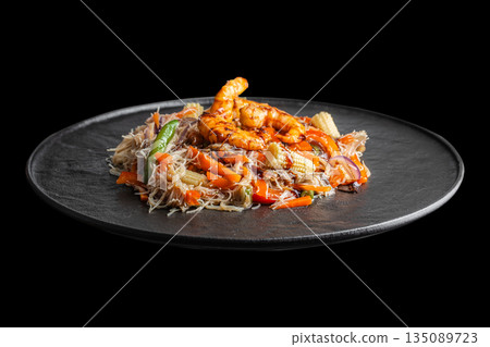 A colorful serving of stir-fried rice noodles with shrimp and mixed vegetables is presented on a black plate against a dark background, showcasing vibrant textures and Asian-inspired flavors 135089723