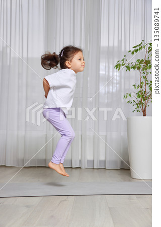 Young girl actively jumping and exercising on a yoga mat in a bright home environment 135089741