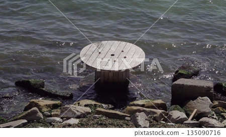 Large wooden cable spool lying in shallow water on the shore, representing environmental pollution, coastal waste and ecological issues affecting marine and shoreline areas Large wooden cable spool lying in shallow water on the shore, representing environmental pollution, coastal waste and ecological issues affecting marine and shoreline areas 135090716