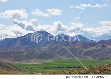 Vast mountain landscape with a green valley and distant snow-capped peaks Vast mountain landscape with a green valley and distant snow-capped peaks 135090782