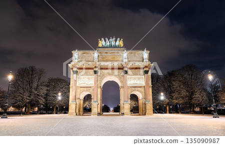 Arc de Triomphe du Carrousel stands near the Louvre in Paris, France. Winter night scene features the illuminated monument and snow-covered grounds with a Ferris wheel in the distance 135090987