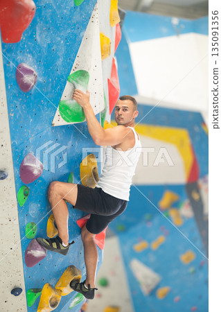 Atlete man grabbing ledges of artificial climbing wall in bouldering centre Atlete man grabbing ledges of artificial climbing wall in bouldering centre 135091356