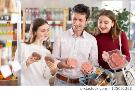 Dad, mom and daughter choose raw chilling beef entrecote and semi-finished hamburger cutlet in shop 135091398